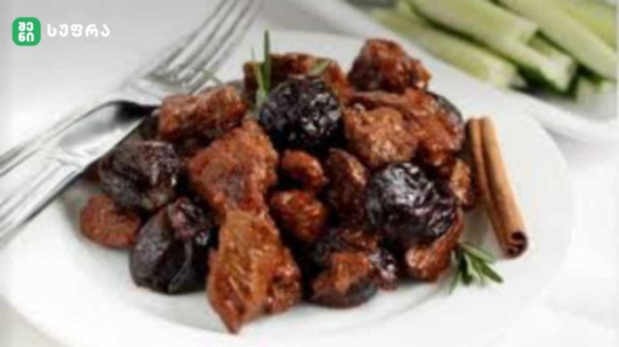 Plate of browned meat chunks with dark prunes, a cinnamon stick, and herbs on a white plate with a fork nearby.