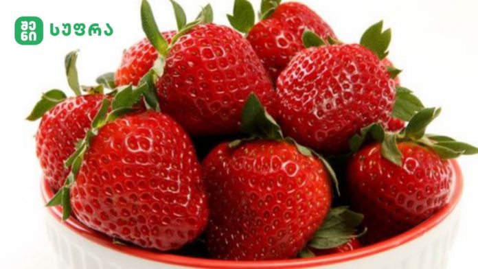 Fresh, ripe strawberries with green leaves in a red bowl on a white background; small green logo with Georgian text in the top-left corner.