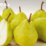 Group of ripe green pears on a light wooden surface, with one pear cut in half showing white flesh.