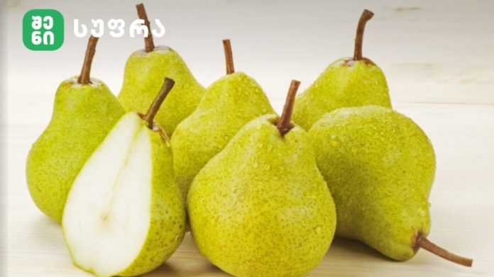 Group of ripe green pears on a light wooden surface, with one pear cut in half showing white flesh.