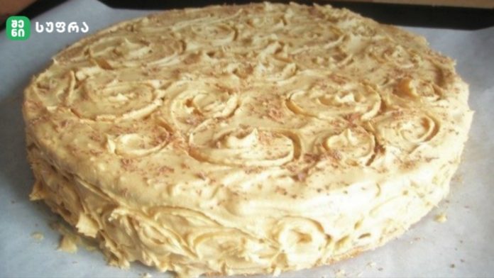 Round vanilla cake with white buttercream frosting and decorative swirl rosettes on top, sides covered in creamy frosting curls, placed on parchment paper.