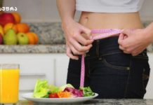 რითი ჩავანაცვლოთ პური? და როგორ დავიკლოთ ზამთრში დაგროვილი ზედმეტი კილოგრამები? Person in a kitchen measures their waist with a pink measuring tape while a plate of salad and a glass of orange juice sit on the counter nearby.