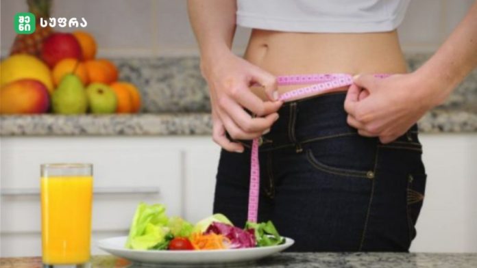 Person in a kitchen measures their waist with a pink measuring tape while a plate of salad and a glass of orange juice sit on the counter nearby.