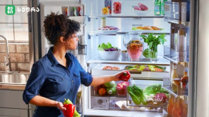 Woman in a blue shirt standing by an open refrigerator, selecting a red bell pepper from fresh vegetables inside.