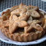 Pie topped with leaf-shaped crust lattice on a white plate, set on a woven table.