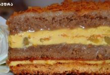 უგემრიელესი კარამელის ტორტი “ჩემპიონი” Close-up of a multi-layer cake with creamy yellow filling, brown cake layers, and a decorative orange topping, with a white flower garnish on the side.