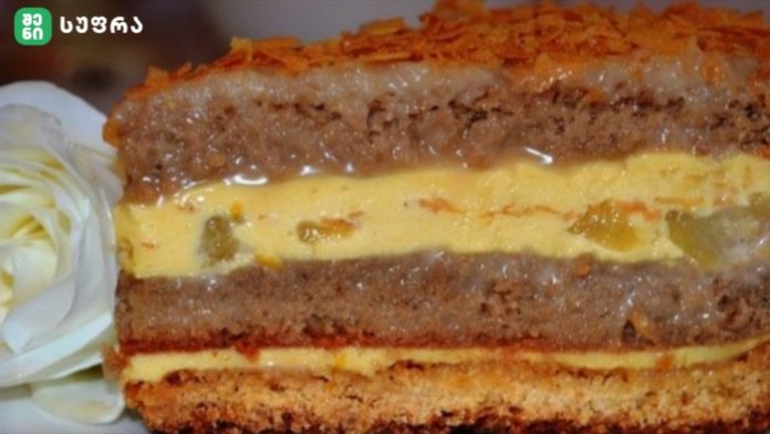 Close-up of a multi-layer cake with creamy yellow filling, brown cake layers, and a decorative orange topping, with a white flower garnish on the side.
