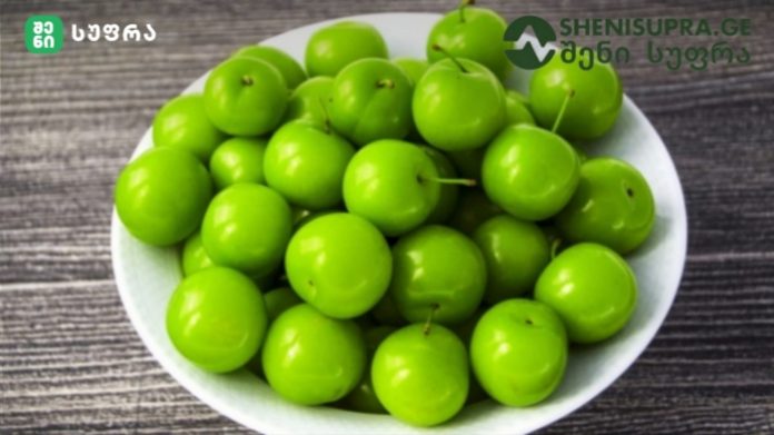 White bowl overflowing with glossy bright green plums on a dark wooden surface.