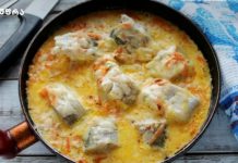 Მინტაი არაჟნის სოუსში Skillet of creamy fish stew with white fish chunks and vegetables in a yellow sauce, cooking on a wooden table.