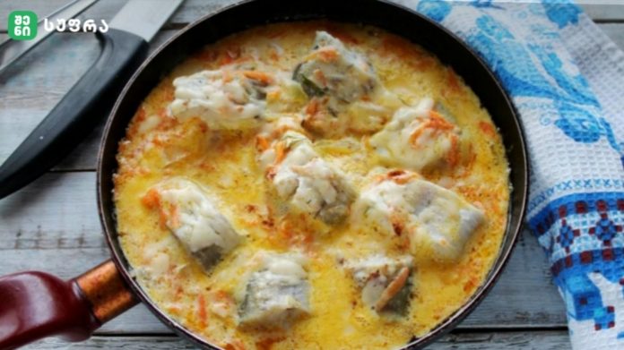 Skillet of creamy fish stew with white fish chunks and vegetables in a yellow sauce, cooking on a wooden table.