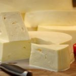 Block of pale cheese with a triangular wedge on a wooden cutting board, knife nearby, plus garlic cloves, cherry tomatoes, bread slices, and a glass of milk in the background.