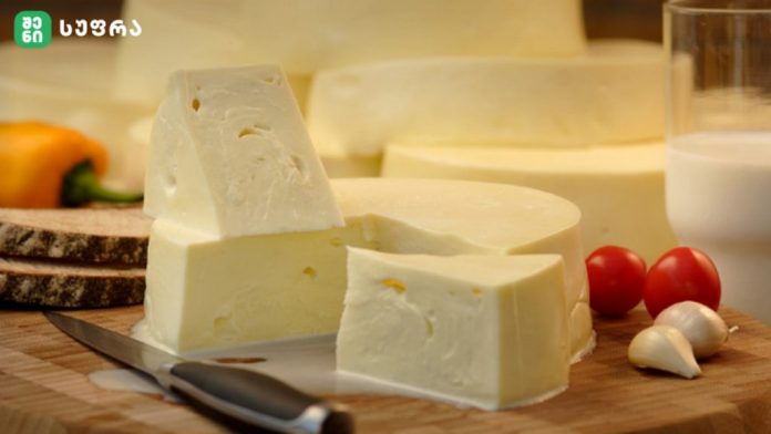 Block of pale cheese with a triangular wedge on a wooden cutting board, knife nearby, plus garlic cloves, cherry tomatoes, bread slices, and a glass of milk in the background.