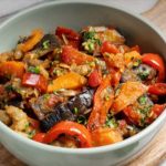 Colorful vegetable medley in a light green bowl: eggplant, red peppers, carrots, and tomatoes with herbs on a wooden board.