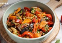 Ბოსტნეულის რაგუ – აჯაფსანდალი Colorful vegetable medley in a light green bowl: eggplant, red peppers, carrots, and tomatoes with herbs on a wooden board.