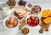 რა უნდა ვიცოდეთ კვებითი ალერგიის შესახებ Assorted breakfast items on a white wooden table: eggs, milk, toast, strawberries, orange slices, nuts, and chocolate pieces.