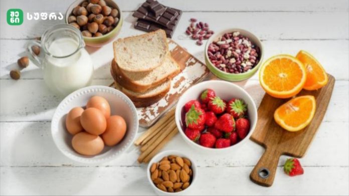 Assorted breakfast items on a white wooden table: eggs, milk, toast, strawberries, orange slices, nuts, and chocolate pieces.