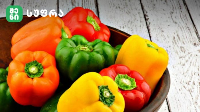 Colorful bell peppers (orange, green, red, yellow) in a bowl on a light wooden table with a green logo in the corner.