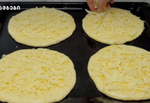 ღუმელში გამომცხვარი ყველიანი ფირფიტები Four unbaked pizza dough rounds topped with shredded cheese on a baking sheet; a hand is sprinkling cheese on one of them.