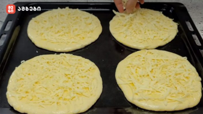 Four unbaked pizza dough rounds topped with shredded cheese on a baking sheet; a hand is sprinkling cheese on one of them.