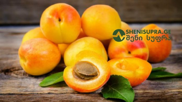 Ripe apricots on a wooden surface, one halved to show the pit, with a green leaf and a SHENISUPRA.GE watermark.