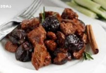 ღორის ხორცი სუნელებით Plate of fried meat chunks with dark prunes, garnished with a cinnamon stick and rosemary on a white plate.
