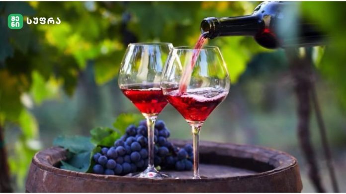 Two glasses of red wine on a wooden barrel as red wine is poured from a bottle, with grape clusters in the background in a vineyard.