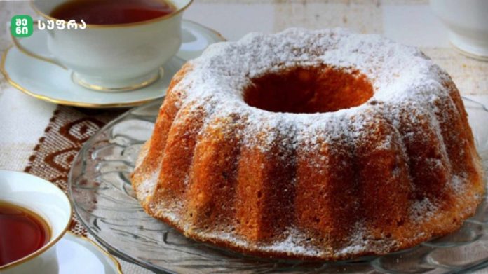 Bundt cake dusted with powdered sugar on a glass platter, with teacups nearby on a table.
