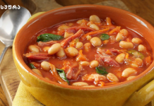ლობიოს სუფი Yellow ceramic bowl of tomato-based white bean soup with herbs on a wooden table, spoon nearby.