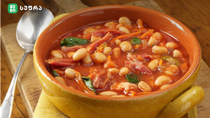 Yellow ceramic bowl of tomato-based white bean soup with herbs on a wooden table, spoon nearby.