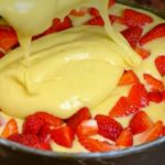 Round springform pan with yellow batter being poured over fresh strawberry pieces arranged around the edges.