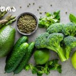 Მწვანე ფერის სარგებელი კვებაში Assortment of green fruits and vegetables on a gray surface: spinach, avocado, cucumber, broccoli, peas, asparagus, lime, and a green apple, with a bowl of peppercorns.