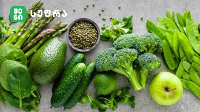Assortment of green fruits and vegetables on a gray surface: spinach, avocado, cucumber, broccoli, peas, asparagus, lime, and a green apple, with a bowl of peppercorns.