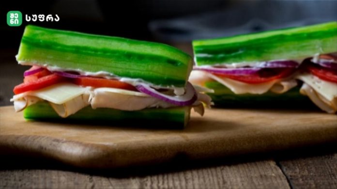Two cucumber sandwiches with layered fillings of cheese, red onion, tomato and greens on a wooden cutting board.