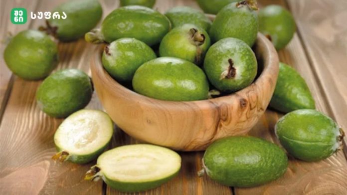 Green feijoa fruits in a wooden bowl on a wooden surface, with a couple cut open to show pale flesh inside.