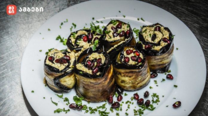 Five rolled eggplant slices filled with a beige stuffing, topped with pomegranate seeds, arranged on a white plate with green herbs.