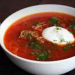 White bowl of red beet soup topped with a dollop of sour cream and chopped herbs, on a dark table with a garlic bulb nearby.
