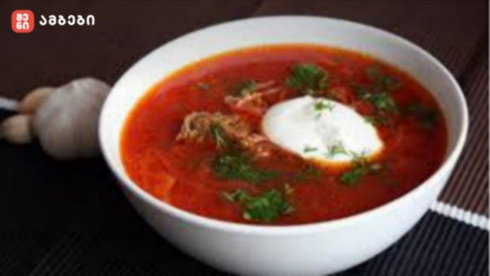 White bowl of red beet soup topped with a dollop of sour cream and chopped herbs, on a dark table with a garlic bulb nearby.