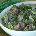 Bowl of beef stew with onions, peppers and herbs on a wooden table, with fresh greens beside it.