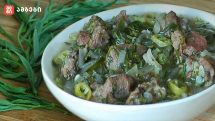 Bowl of beef stew with onions, peppers and herbs on a wooden table, with fresh greens beside it.