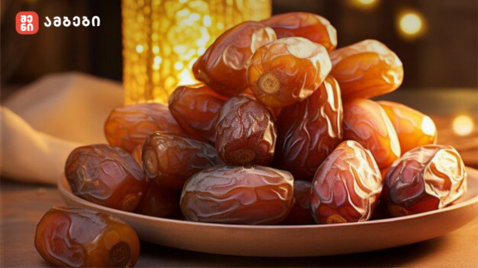 Pile of glossy dates on a light plate under warm ambient lighting; logo in the top-left corner.