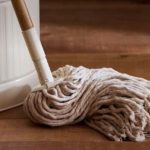 Beige mop head wringing on a wooden floor beside a white bucket.