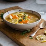 Bowl of lemon-garnished vegetable soup on a wooden cutting board with lemon slices and a spoon nearby.