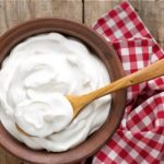Bowl of creamy yogurt with a wooden spoon on a rustic wooden table, with a red‑and‑white checkered cloth nearby.