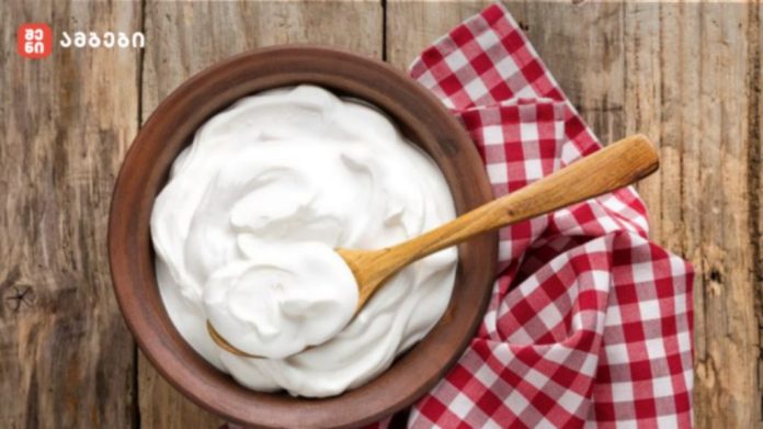 Bowl of creamy yogurt with a wooden spoon on a rustic wooden table, with a red‑and‑white checkered cloth nearby.