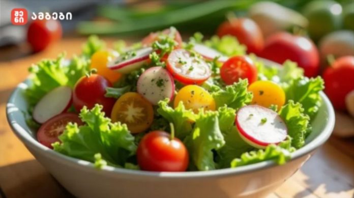 White bowl of mixed greens salad with lettuce, cherry tomatoes, and radish slices on a wooden table; logo visible in the top-left corner.