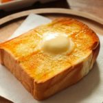 Square slice of toasted bread with a knob of butter melting on top, placed on a white napkin on a wooden plate.