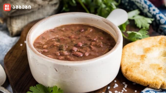 Bowl of hearty bean soup in a white ceramic bowl on a wooden board, with crusty bread and fresh herbs nearby.