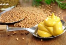 როგორ მოვამზადოთ მდოგვი სახლის პირობებში A spoon filled with swirled yellow mustard on a wooden surface, with mustard seeds scattered nearby and green herbs blurred in the background