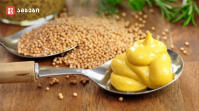 A spoon filled with swirled yellow mustard on a wooden surface, with mustard seeds scattered nearby and green herbs blurred in the background