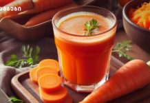 სტაფილოს ფრეში -ვიტამინების საბადო Glass of fresh carrot juice on a wooden board with sliced carrots and a whole carrot nearby, garnished with a herb sprig.